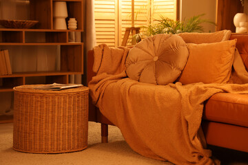 Cozy sofa with plaid, cushions and wicker basket in interior of living room