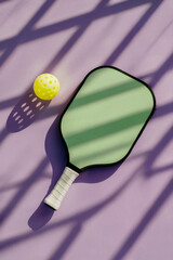 Modern pickleball paddle and yellow perforated ball on pastel purple surface with dramatic fence shadow, summer sport concept, vertical composition for editorial or lifestyle use