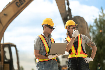 Two construction workers in hard hats and reflective vests discussing blueprints at an industrial...