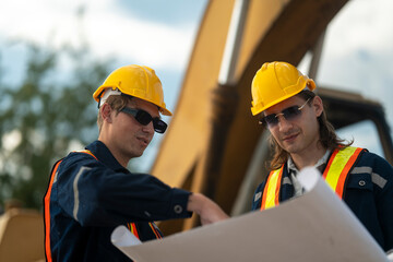 Two construction workers in hard hats and reflective vests discussing blueprints at an industrial site, emphasizing collaboration, planning, and safety in civil engineering projects.