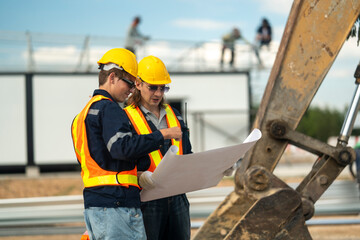 Two construction workers in hard hats and reflective vests discussing blueprints at an industrial...