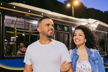 Young Latin woman happily holds her partner's arm to walk after getting off the bus behind them