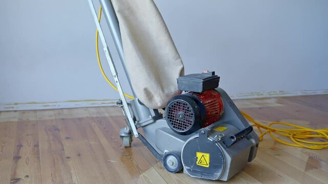 Electric floor sander stands on partially sanded pine wood floor, dust collection bag and power cable attached, positioned indoors against a plain wall, used for professional parquet restoration work.