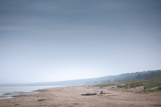 Garciems Beach on Latvia Baltic coast lies deserted in misty rain; pale sand, scattered driftwood and a hazy tree line dissolve into low clouds, conveying the tranquil mood of a cloudy afternoon.