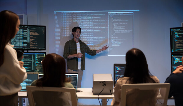 Asian man presents code using a projector during a software development meeting or coding workshop, guiding attendees through problem-solving and programming concepts in an educational setting.