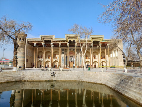 The magnificent Bolo Haouz Mosque in Bukhara, Uzbekistan, reflected in its tranquil reflecting pool. The mosque's facade features a long colonnade of slender, intricately carved wooden columns