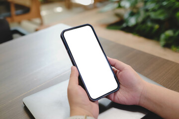 Person holding smartphone with blank screen at wooden table in cafe, resting hand on closed laptop, showing technology in relaxed environment.