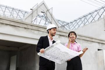 Two architects wearing safety helmets discuss blueprints at a construction site. Surrounded by scaffolding and a partially built structure, they collaborate on the details of the building project.