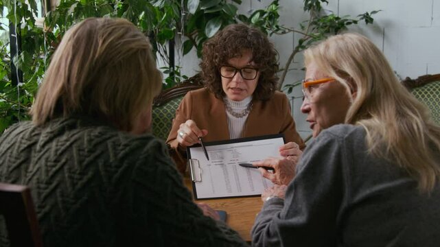 Medium shot of cheerful Caucasian female mortgage broker consulting couple of mature clients considering taking bank loan, explaining terms and rates, demonstrating loan amortization schedule
