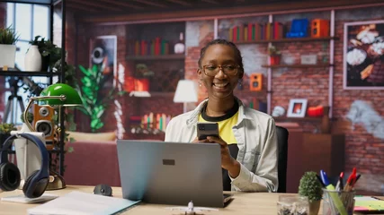 Selbstklebende Fototapeten Portrait of jolly woman sitting on chair at home and texting friends on smartphone. Cheerful african american person in living room enjoying leisure time, chatting with mates, camera B © DC Studio
