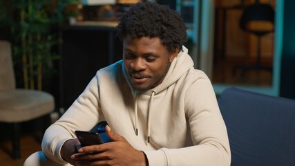 African American man on a sofa texting mates in his living room, entertained by social media and messages. Depiction of remote work breaks, digital communication and relaxation at home. Camera A.