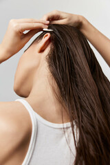Fototapeta premium Woman styling her long hair, with her back to the camera, displaying glossy strands against a minimalistic light background Emphasizes beauty and personal care