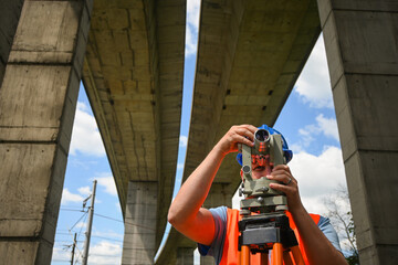 Surveyor taking measurements with theodolite underneath concrete highway overpass pillars