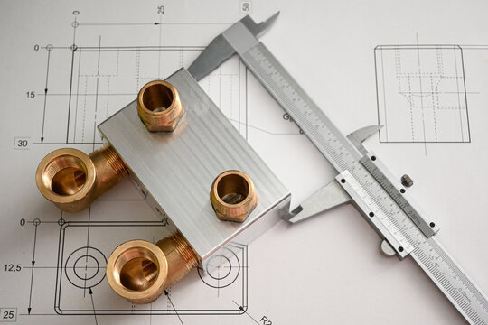 Metal plumbing fixture with brass connectors lying on a blueprint is being measured with a caliper