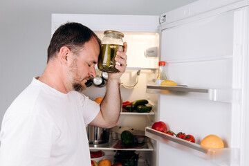 Man holding jar of pickles against forehead, standing in front of open refrigerator filled with...