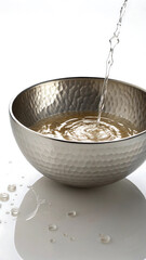 Water pouring into a hammered metal bowl creating ripples and water droplets on a reflective surface.