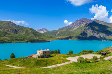 Alpine landscape with a vibrant turquoise lake of Mont Cenis, rocky mountains under a bright blue sky with white clouds in France.