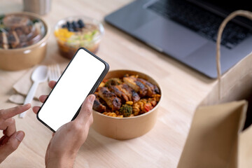 Hand holding a smartphone with blank white screen over paper bowl with healthy food, Young man, woman eating healthy meal while working, Businessperson having a lunch break at the office or home.