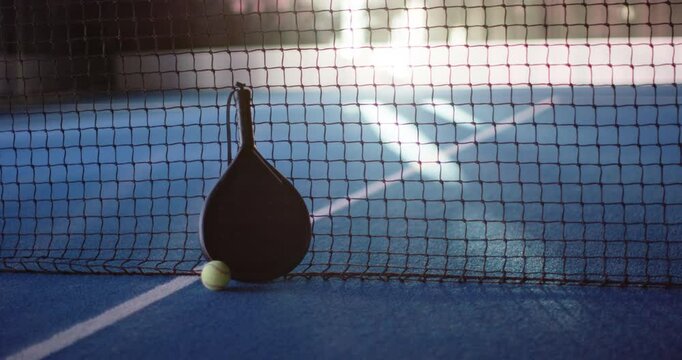 Padel racket and ball resting against net on blue court, copy space