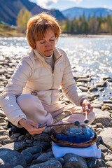 Woman playing a steel tongue drum near a mountain river in Altai, surrounded by wild nature. Concept of spiritual retreat, sound healing, and ancient connection to earth