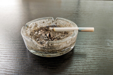 Dirty glass ashtray with ashes and cigarette butts on dark wood table, one partially smoked cigarette on edge. Concept of addiction, smoking habit, and health risk.