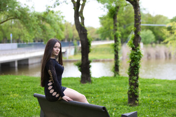 Obraz premium Latin woman, young and beautiful brunette dressed in black sitting on the back of a park bench looks mysteriously and sensually backwards. In the background a lake on a gray day.