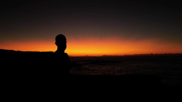 Sillhouette of man wearing baseball cap looking out to the ocean at sunrise