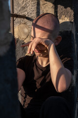 Vertical photo of a shaved-head teenage boy in a prison cell, sitting by a barred window and wiping tears from his face. A young inmate crying helplessly fear, despair, and lost childhood.
