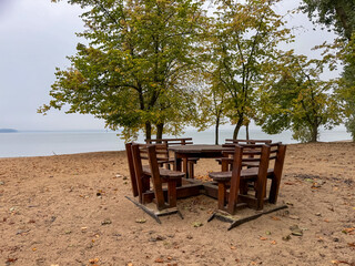 Obraz premium Wooden round table and chairs on a deserted lakeside beach under an overcast sky, peaceful and moody scene