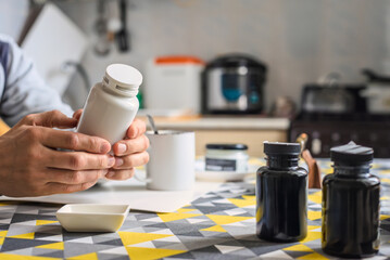 Closeup of male hands holding bottle with natural vitamins at home kitchen. Man sitting at kitchen table holding supplement bottle with other jars of dietary additives nearby.