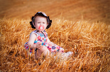 Adorable baby girl in a colorful dress and sunhat sits in a golden wheat field with a pacifier. Captures the essence of rural childhood in warm summer light
