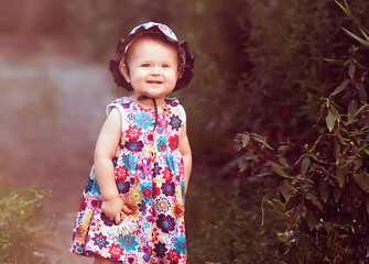 Smiling toddler girl in a colorful floral dress and sunhat stands outdoors with a daisy in hand. Joyful childhood moment in natural summer light