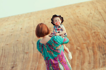 Mother holding her baby daughter in arms, standing outdoors against a sandy hill. Family love, summer bonding, and parenthood concept in natural light