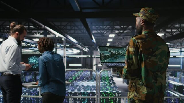 Military technician reviewing tactical dashboard on laptop screen inside secure server hub. Army expert using strategic communications and infrastructure oversight software on notebook, camera A