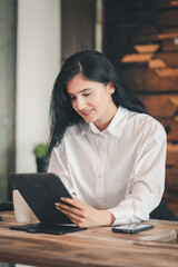 Fototapeta premium Hispanic woman in white blouse holding coffee cup, smiling at tablet screen in office. Modern workspace with computer monitor and decor. Relaxed professional setting, positive workplace atmosphere