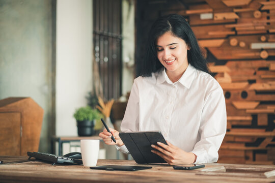 Hispanic freelancer woman working relaxed smiling looking at tablet in office. graphic designer female working on computer and looking monitor, professional creative occupation at workplace lifestyle