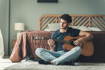 Male guitarist sitting on carpeted floor with acoustic guitar across legs, practicing quietly in relaxed bedroom space, portraying peaceful creativity and passion for music, man on holiday at home © Rakchanok