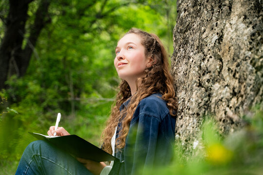 Dreamy young woman with curly hair writing something in notebook outdoors looking up relaxing resting alone keeping diary journaling dressed casual dark blue shirt sitting in nature park, portrait