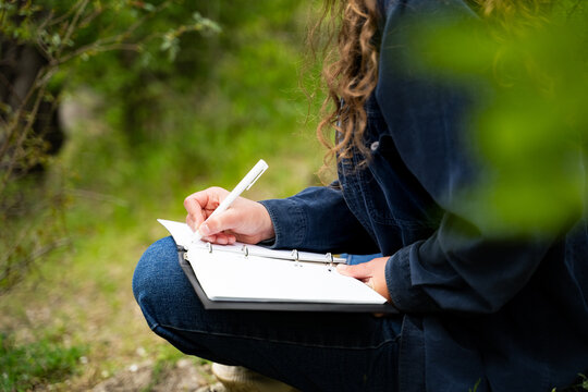 Unrecognizable woman with curly hair writing in notebook outdoors resting relaxing spending leisure time alone keeping diary journaling dressed casual dark blue shirt and jeans sitting in nature