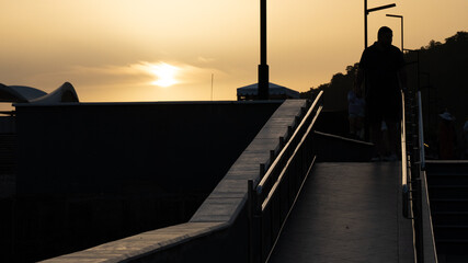 Silhouettes of People Walking at Sunset on a Modern Waterfront Ramp — Urban Evening Scene with Warm Light and Shadows