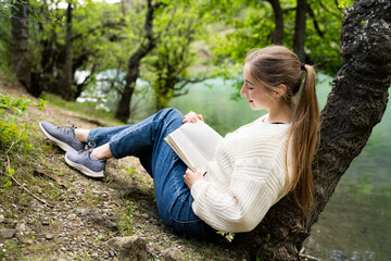 Relaxed happy young woman enjoying reading a book outdoors feeling happiness and calmness wearing...