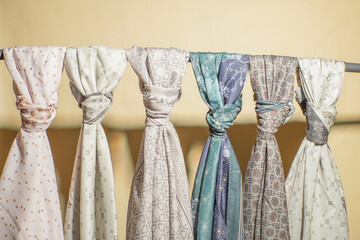 A collection of colorful patterned hijabs neatly tied and hanging on a rod with a soft beige background. The fabrics feature intricate designs, suitable for showcasing Islamic fashion, textiles, or m