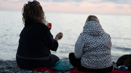 Two Women Sitting on a Pebble Beach at Sunset with Drinks — Relaxing Seaside Moment