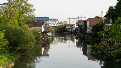 Canal with Stilt Houses and Reflections, Framed by Greenery and Railway Bridge in the Distance