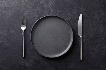 Empty black plate with a fork and knife on a dark stone background. Flat lay. Copy space. Top view.