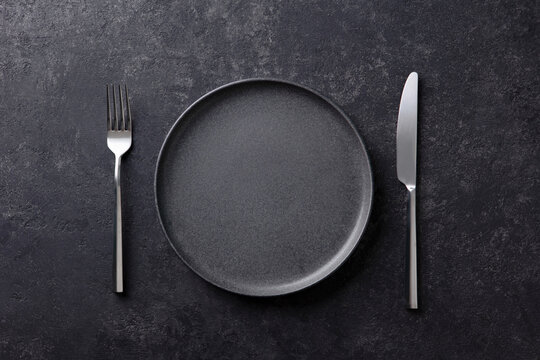 Empty black plate with a fork and knife on a dark stone background. Flat lay. Copy space. Top view.