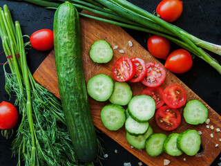 Red cherry tomatoes with herbs and cucumber sprinkled with sea salt on the wooden board on black table, top view of vegetables covered with water drops.