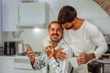 Tattooed gay couple hugging while drinking coffee and using smart phone