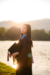 Asian woman graduate, holding her cap, stands by a serene lake with mountains at sunset, symbolizing accomplishment and future aspirations.