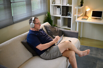 Senior man with glasses and headphones using digital tablet on couch